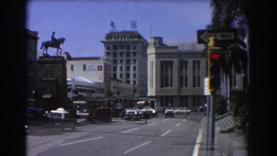 1967 Brisbane urban street scene showing classic cars, a large civic-style building, palms, an equestrian statue, and a red signal.