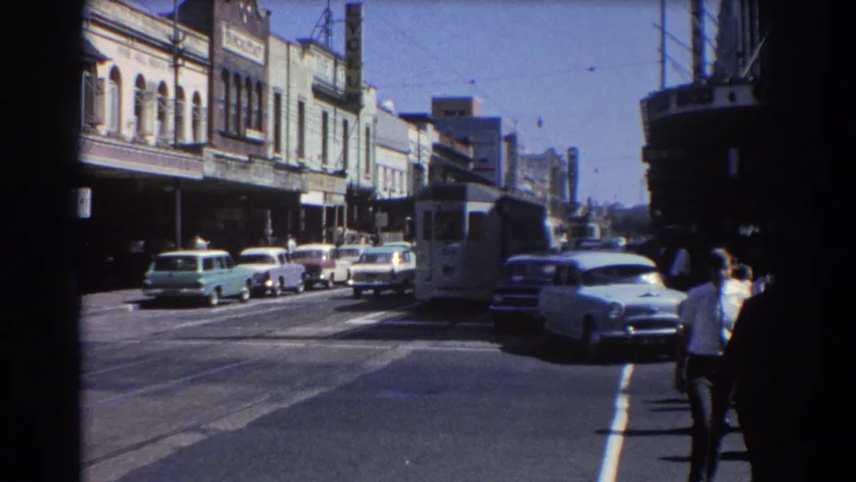 1967 Brisbane street view with storefront facades, parked mid-century cars, overhead wires, a tram or bus-like vehicle, and pedestrians.