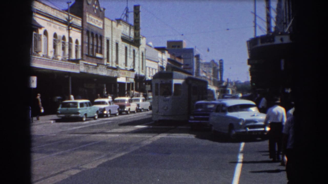 White electric tram approaching on a Brisbane street with cars, shopfronts, and pedestrians nearby.