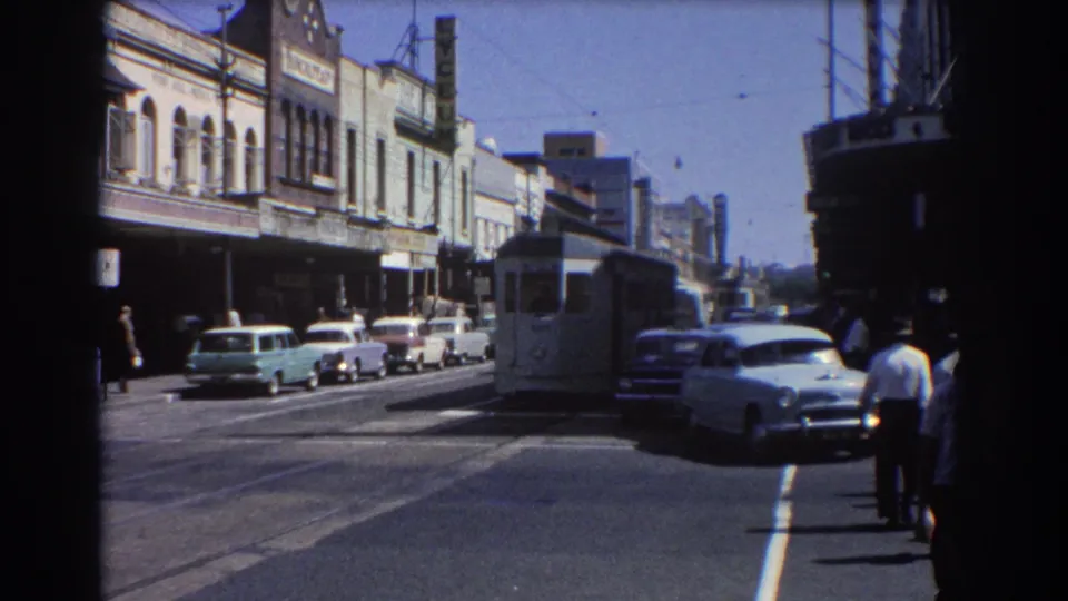 1967 Brisbane street view with a white electric tram in the roadway, mid-century cars, overhead wires, and pedestrians along the sidewalk.