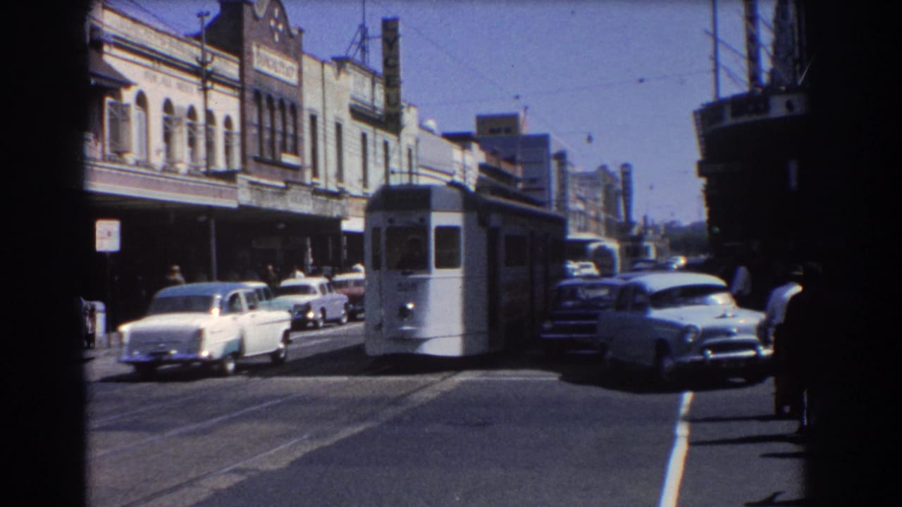 White tram and classic cars move through a sunlit street lined with older commercial buildings.