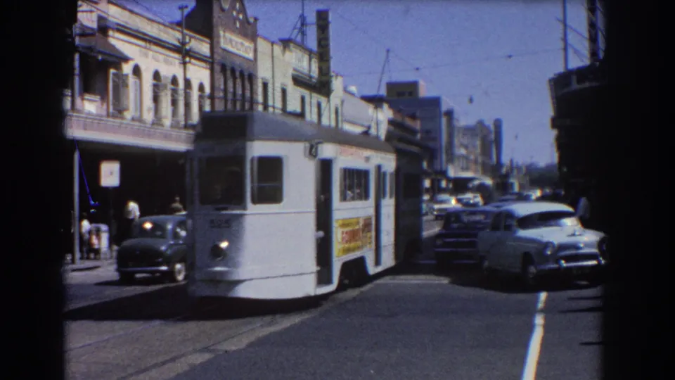 A white electric tram passes close to the camera in Brisbane, with side advertising, classic cars, shopfronts, and pedestrians in view.