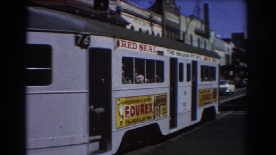 Close side view of a Brisbane white electric tram showing Red Seal and Fourex ads, passenger windows, and cars behind it.