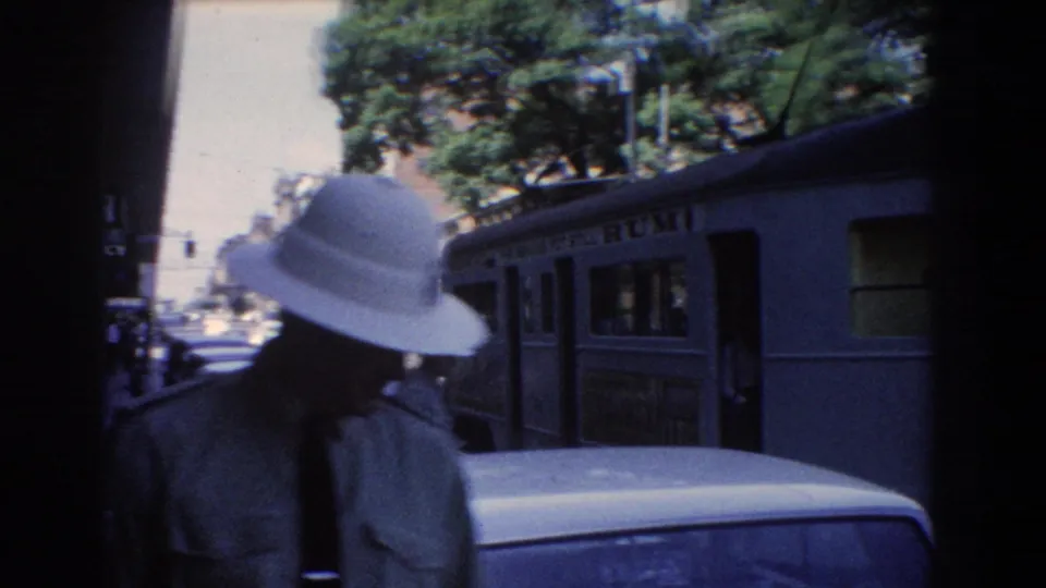 1967 Brisbane street scene with a tram partly in shade, older cars, shopfronts, and a pedestrian in a white hat near the foreground.