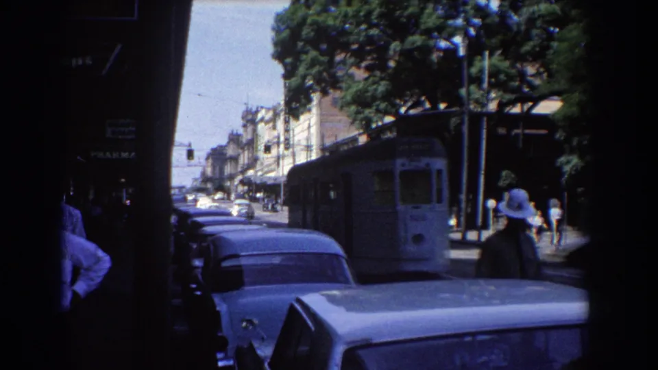 1967 Brisbane street view with a tram receding along the road, older cars in the foreground, pedestrians, trees, and shaded storefronts.