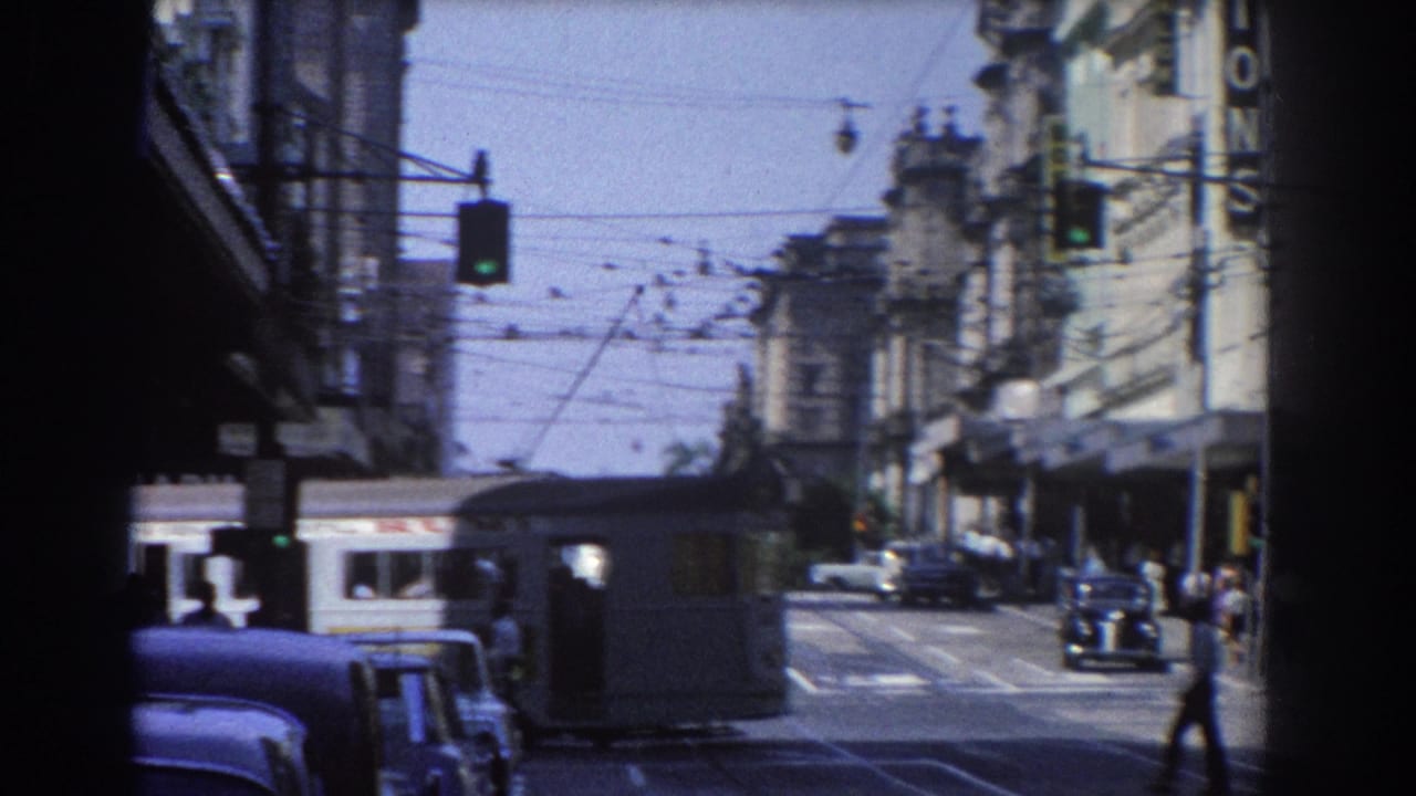 1967 Brisbane street view with vintage cars, a trolley car, pedestrians, traffic lights, and overhead trolley wires framed by a dark foreground edge.