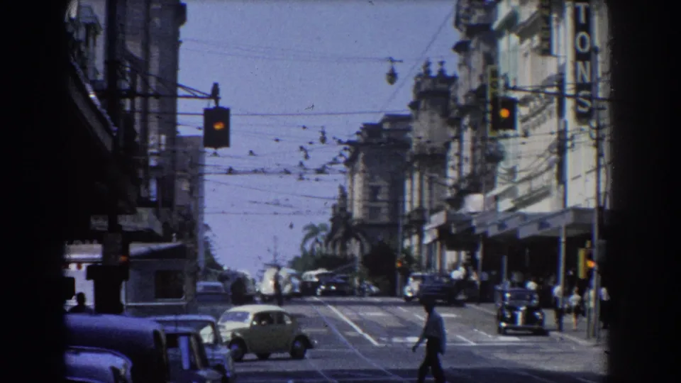 A Brisbane intersection in 1967 with a small car crossing, pedestrians near storefronts, traffic lights, overhead wires, and older automobiles.