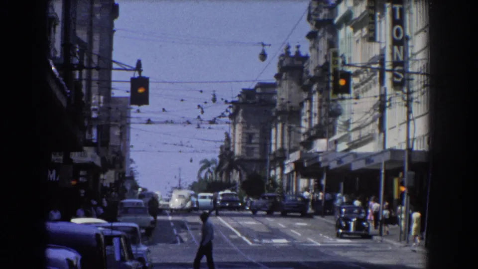 1967 Brisbane street traffic with a pedestrian crossing, vintage cars in both directions, overhead trolley wires, and storefronts along the road.