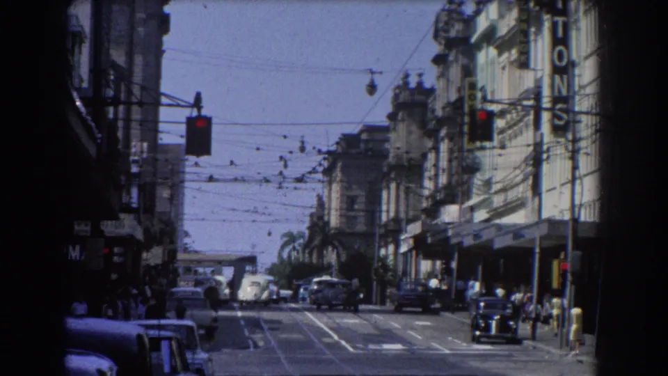 A 1967 Brisbane street view with cars moving under trolley wires, pedestrians near shopfronts, traffic lights, and bright mid-day exposure.