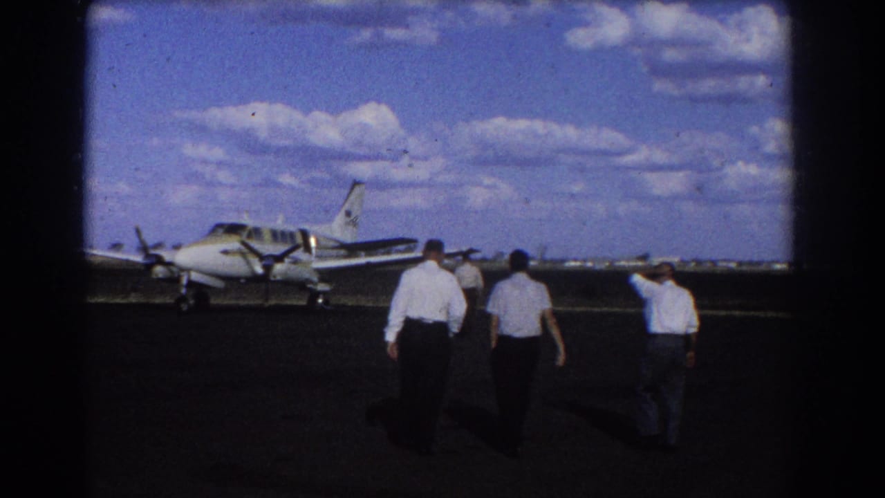 Three men walk across a dark tarmac toward a small twin-engine propeller plane under a blue sky.