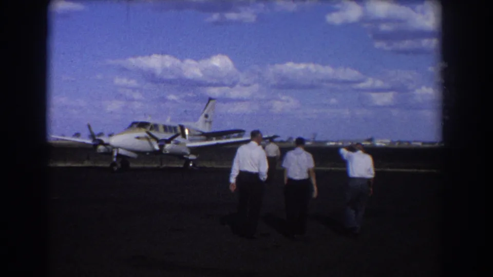 Three men walk farther across the Brisbane airport tarmac toward a small twin-engine aircraft in 1967 home-movie footage.