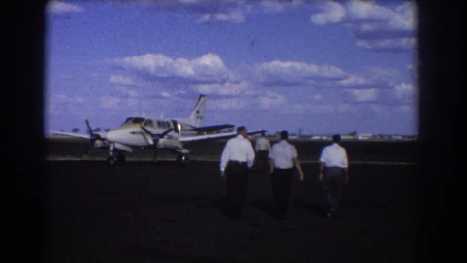 In the 1967 Brisbane airport scene, three men in shirts and trousers approach the side of a small parked propeller plane.