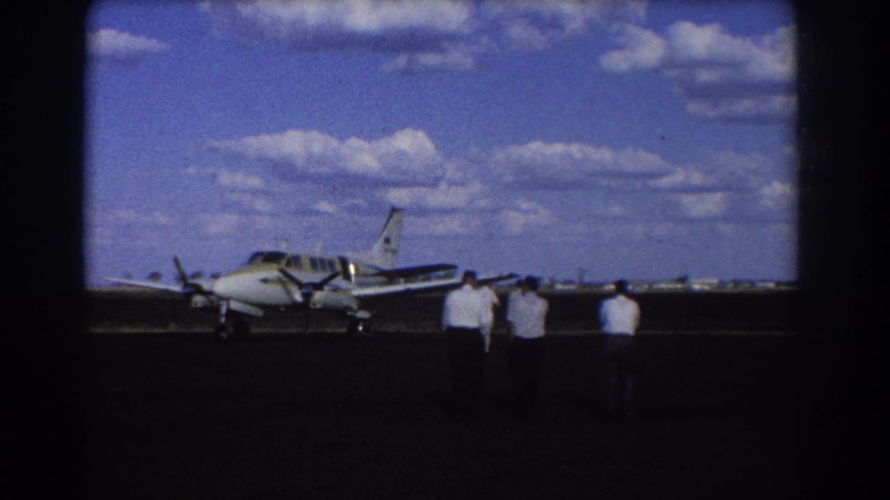 Small propeller plane on a tarmac with three men walking toward it under a blue sky.