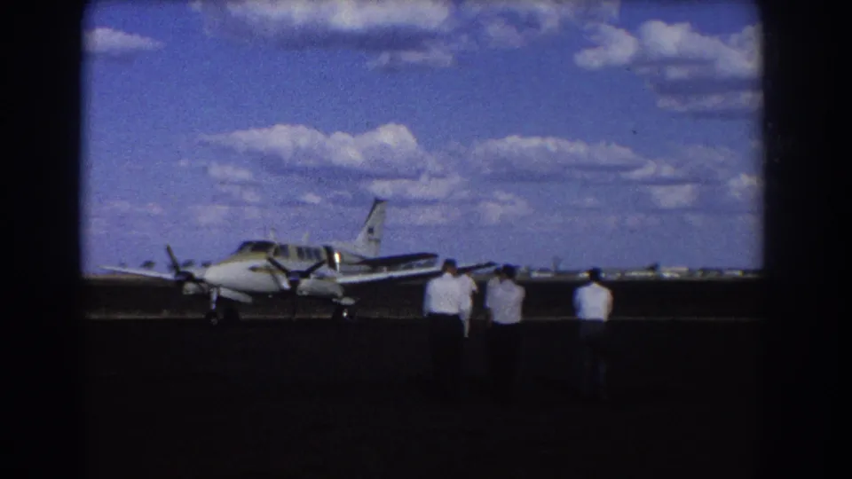 1967 Brisbane 8mm frame showing men in white shirts walking across an airport tarmac toward a small propeller plane.