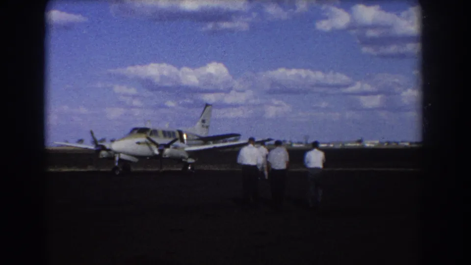 1967 Brisbane home-movie still with several men nearing a small twin-engine plane on a sunlit tarmac.