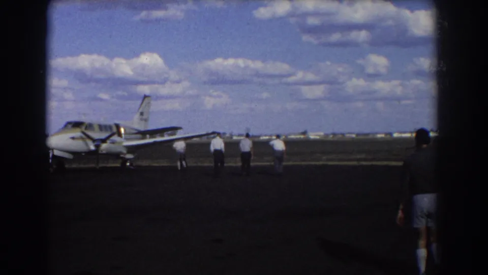 1967 Brisbane 8mm frame showing a small propeller plane, men on the tarmac, and a foreground figure partly in shadow.