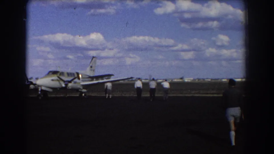 1967 Brisbane home-movie frame of men approaching a small aircraft on a runway, with a shadowed figure at the right edge.