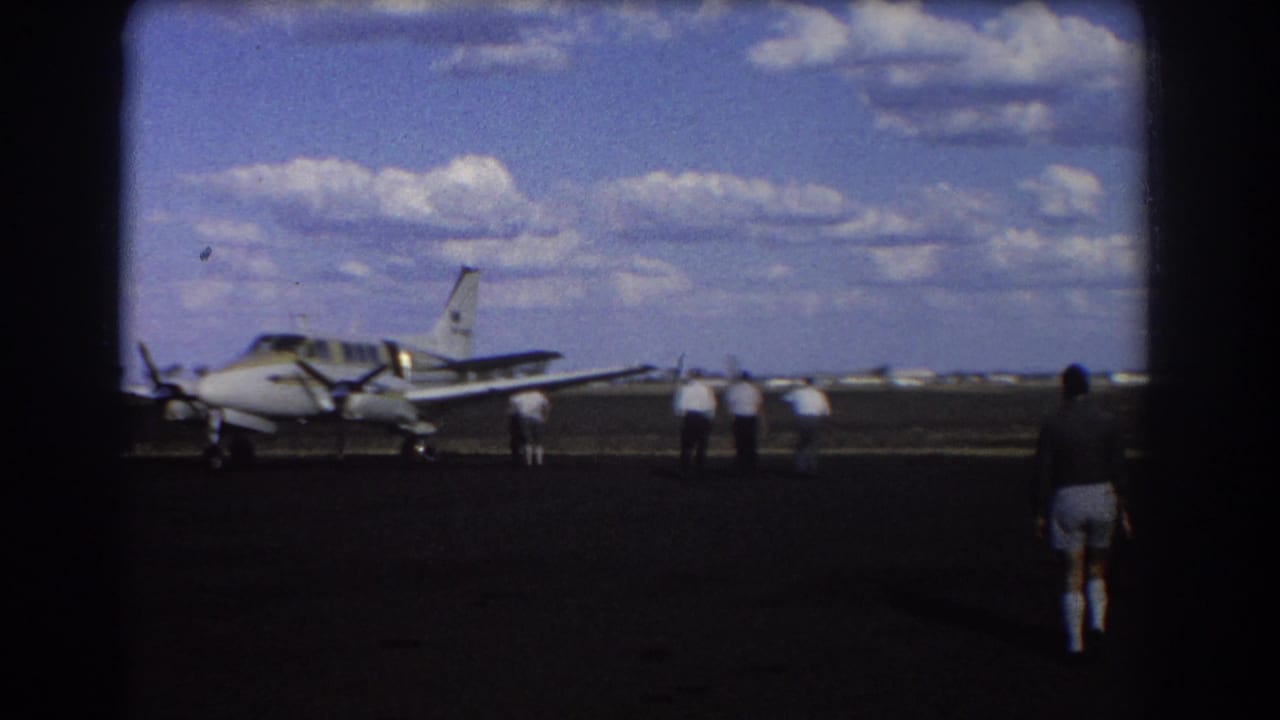Small twin-engine propeller plane parked on a runway with several men nearby under a cloudy sky.