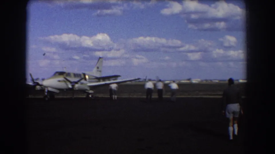 1967 Brisbane home-movie frame showing a small propeller aircraft on tarmac with men standing and walking nearby.