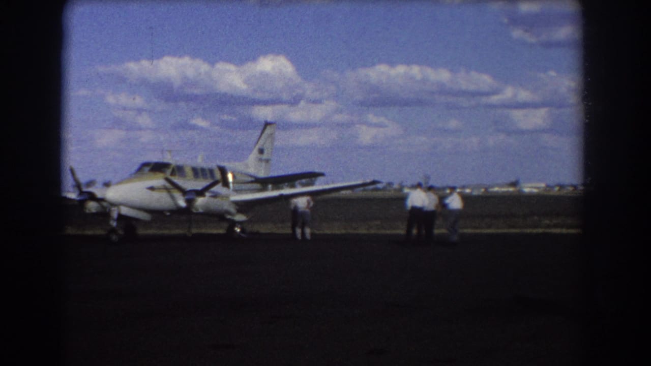 Parked propeller aircraft with men standing near the right side of the runway scene.