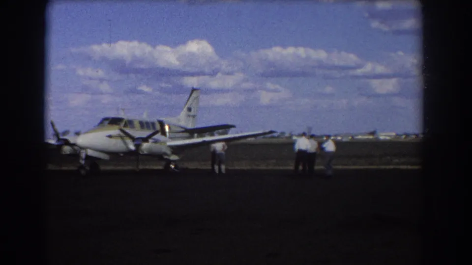 1967 Brisbane 8mm frame of a small aircraft parked on an airfield while men stand near its wing area.