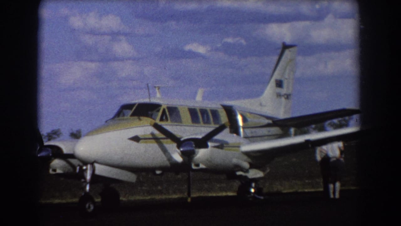1967 Brisbane home-movie frame showing a white and yellow twin-engine propeller airplane parked on a sunlit grass airfield.