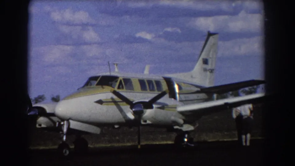 1967 Brisbane home-movie frame showing a white and yellow twin-engine propeller airplane parked on a sunlit grass airfield.