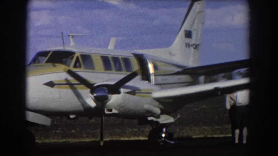 1967 Brisbane frame with the parked twin-engine aircraft closer in view, showing yellow striping, cockpit windows, and tail markings.