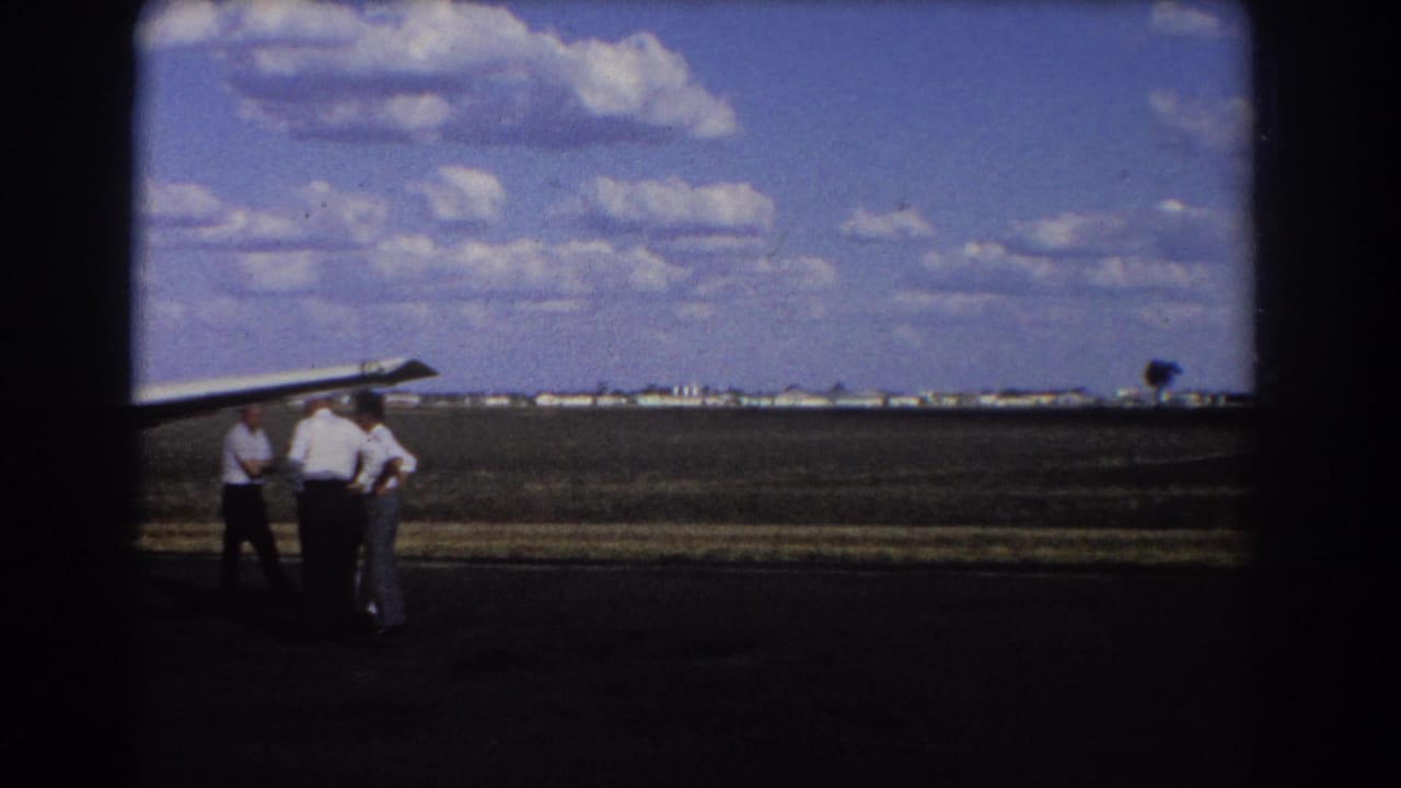 Three men stand near a small aircraft wing on an open airstrip under a cloudy blue sky.