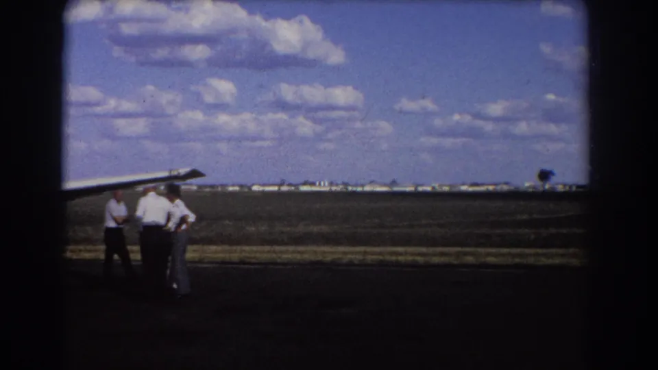 1967 Brisbane home-movie frame showing three men near a small aircraft wing on a wide airstrip with distant buildings.