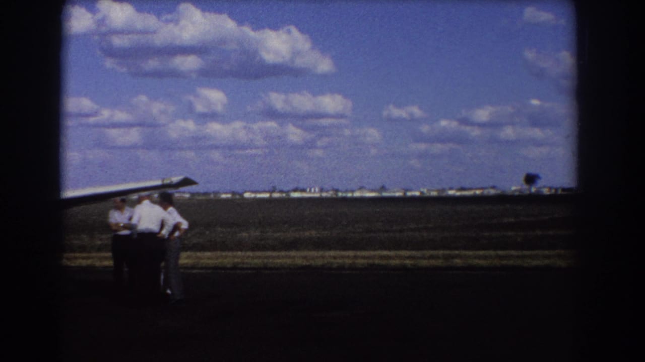 Men in white shirts stand near an aircraft wing with flat airfield land and clouds behind them.