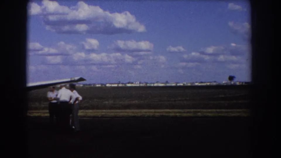 1967 Brisbane airstrip view with several men beside a small aircraft wing and low buildings on the horizon.