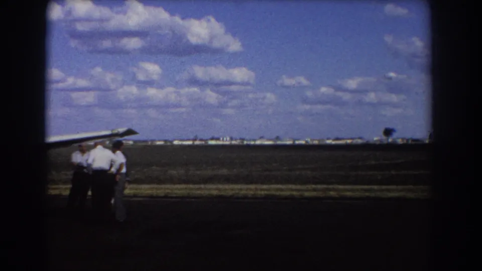 1967 Brisbane home-movie frame with men clustered by a small aircraft wing at the edge of an open airfield.