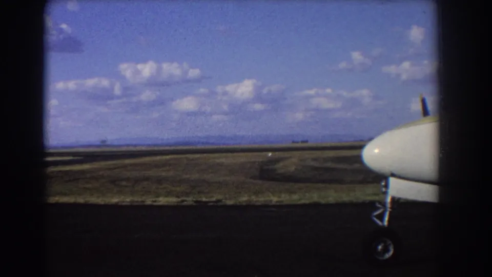 1967 Brisbane airstrip frame showing the front of a small aircraft, landing gear, runway, grassland, and distant horizon.