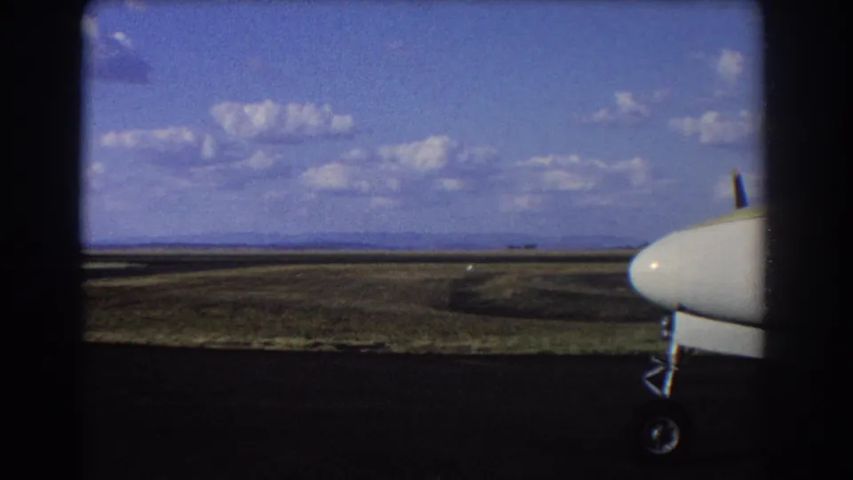 1967 Brisbane airfield view with the nose and landing gear of a small aircraft at frame right and open runway terrain beyond.