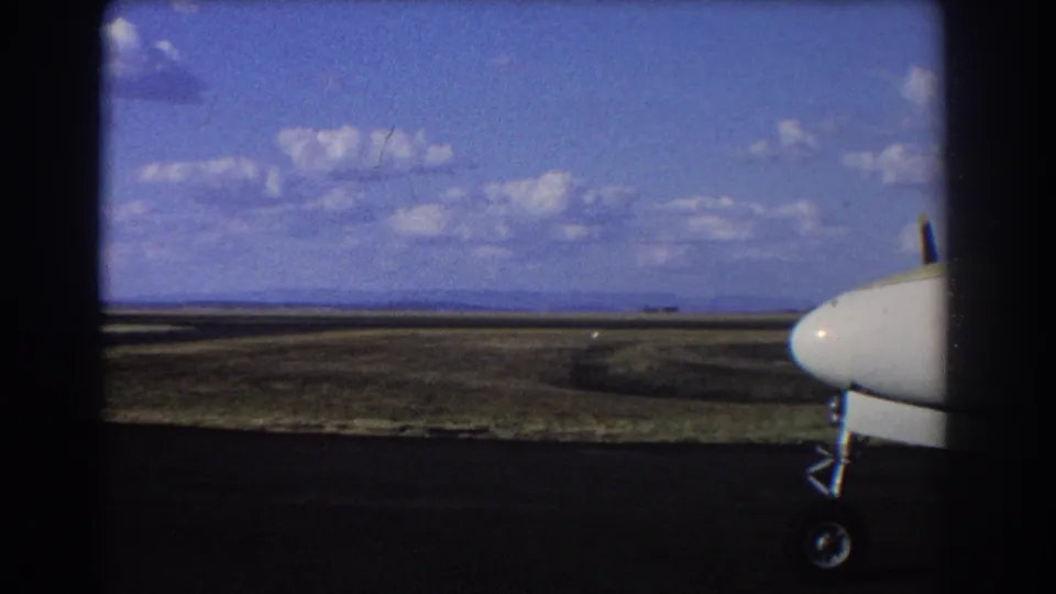 1967 home-movie frame showing a small aircraft at frame right, tarmac in shadow, and open Brisbane-area airfield terrain beyond.
