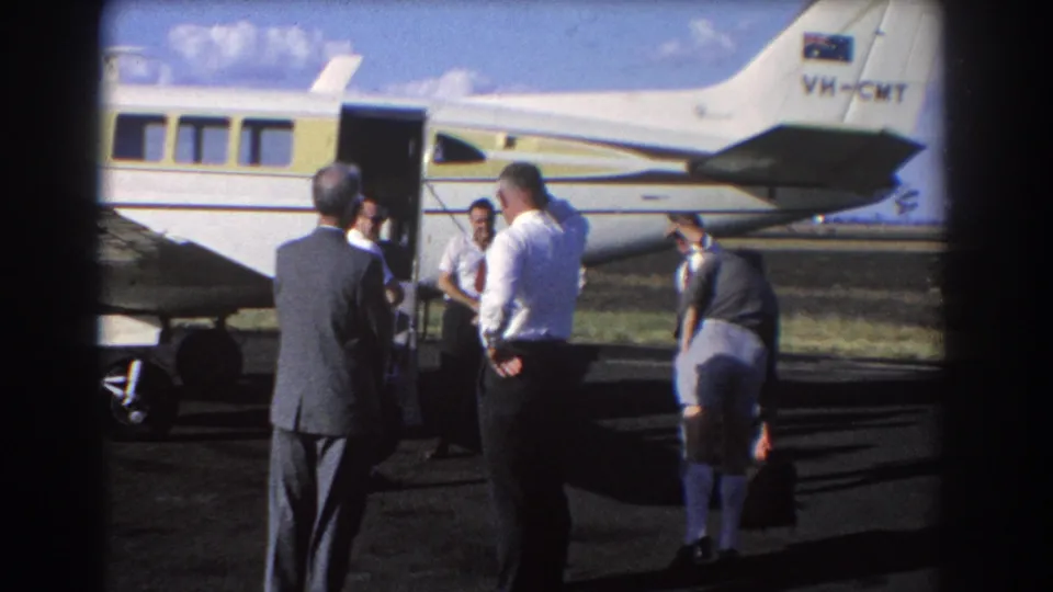 In Brisbane, Australia in 1967, men in suits and shirts stand near the open door of a small white and yellow aircraft on the tarmac.