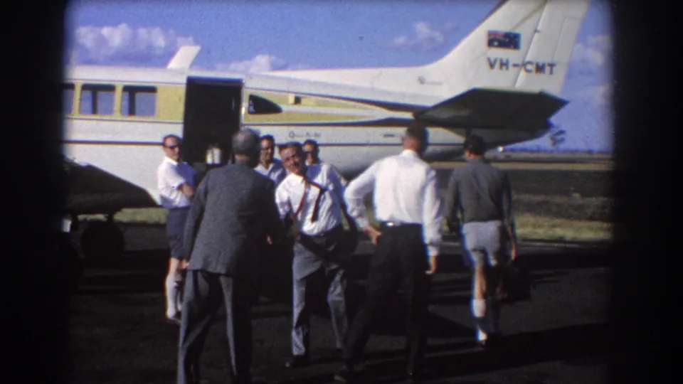 Men gather casually by a small aircraft in Brisbane in 1967, including suited figures and a man in shorts carrying a briefcase.