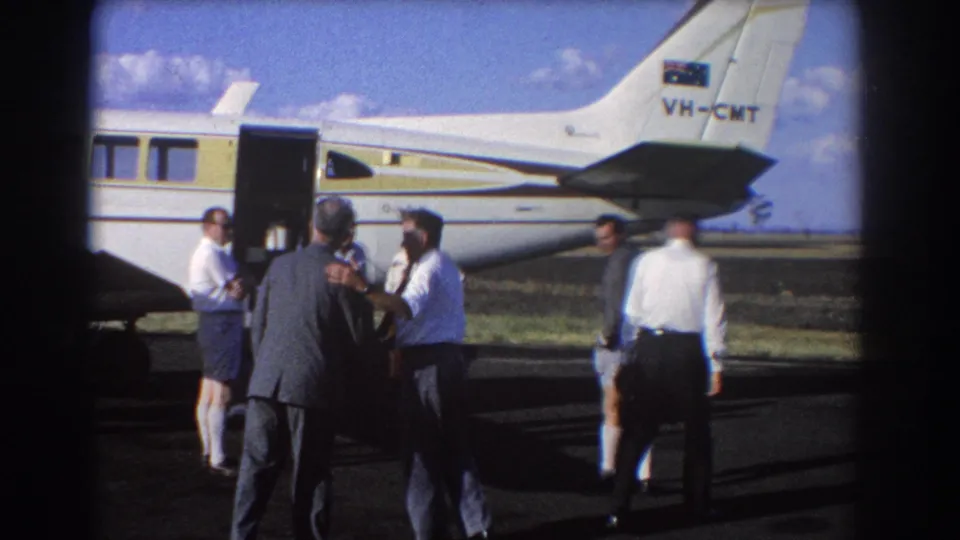 A 1967 Brisbane tarmac scene shows men near the open door of a small aircraft while one man in shorts walks away carrying a briefcase.