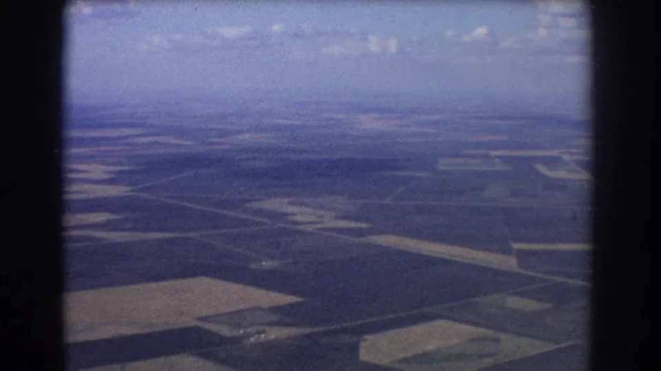 Late-1960s Brisbane-area aerial frame with patchwork fields, road lines, sky, and home-movie film texture.