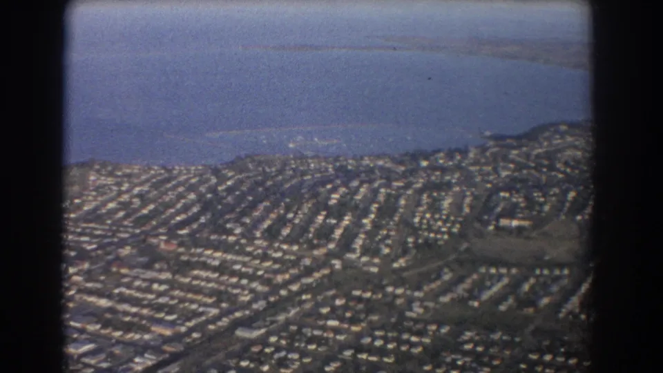 1967 Brisbane-area aerial frame with dense low-rise suburbs, curving streets, hills, and a broad water body beyond.