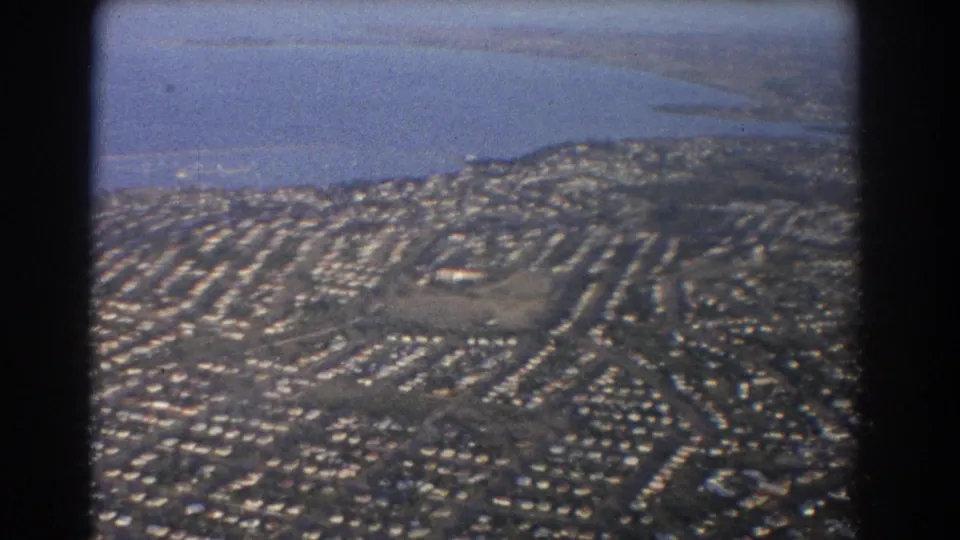 1967 Brisbane-area 8mm frame showing dense residential streets, a larger open site, low hills, and water in the background.