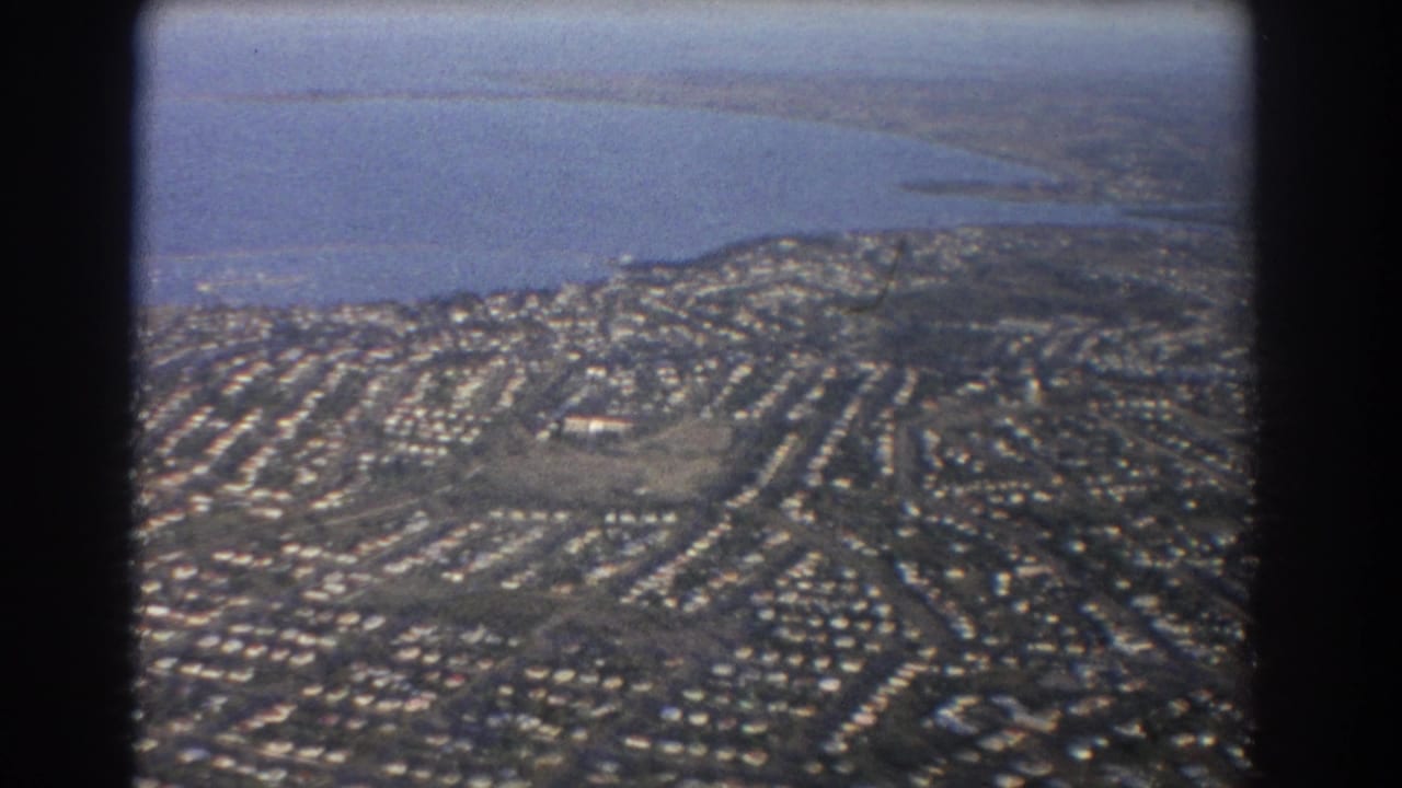 1967 Brisbane-area aerial view showing dense suburban housing, curving streets, and a broad coastal waterline in soft 8mm color.
