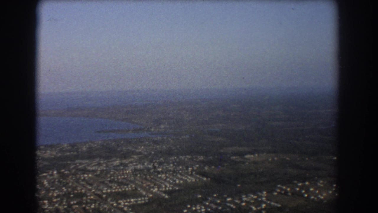 Aerial view over suburbs, open land, and a body of water under a hazy sky.