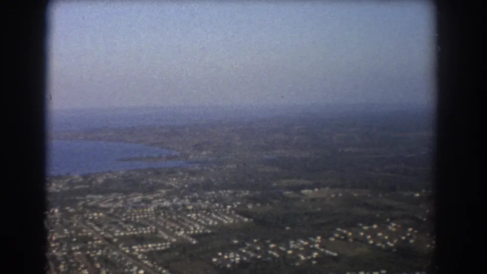 1967 Brisbane-area aerial frame showing shoreline water, patterned suburban streets, scattered houses, and a hazy inland horizon.