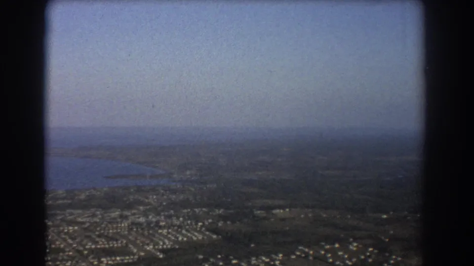1967 Brisbane-area aerial view with a receding shoreline, low suburban housing, open fields, and soft haze across the horizon.