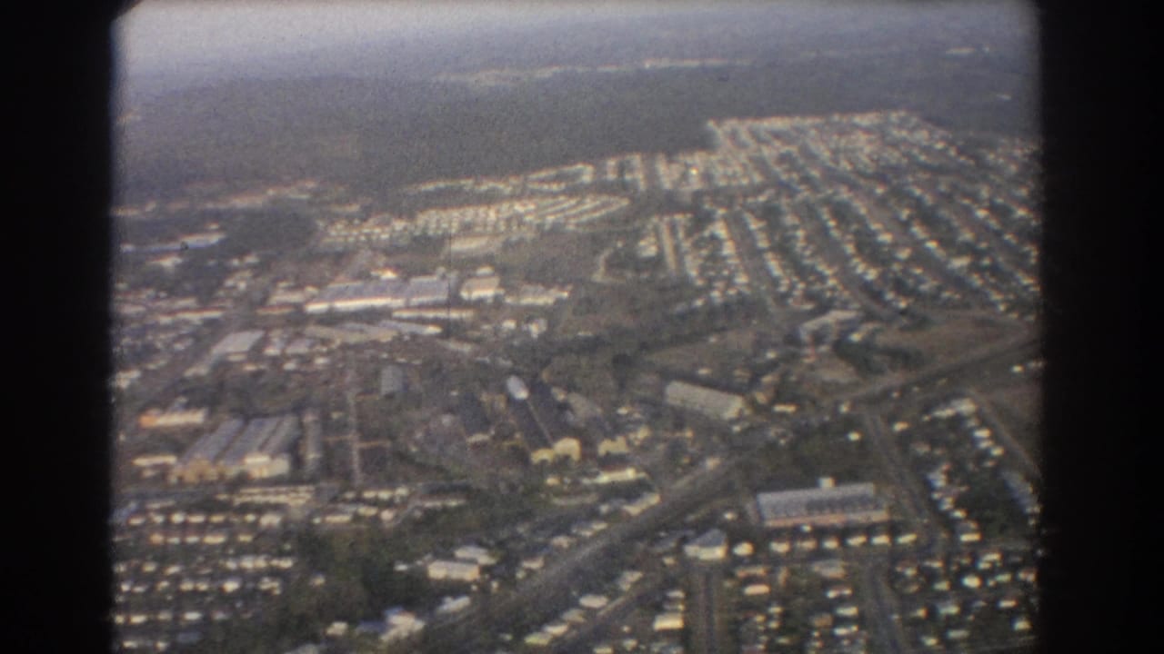 1967 Brisbane aerial footage showing rows of suburban houses, roads, open land, and small larger buildings from a plane.