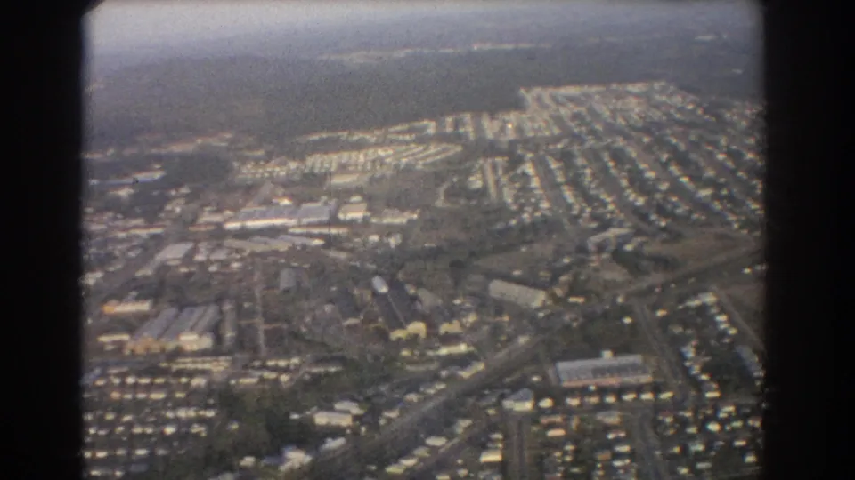 1967 Brisbane aerial footage showing rows of suburban houses, roads, open land, and small larger buildings from a plane.