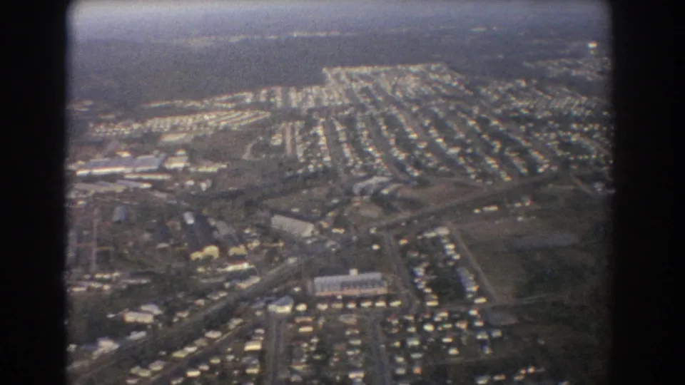 Aerial view over Brisbane in 1967, with housing rows, roadways, open parcels, and distant tree cover visible below.