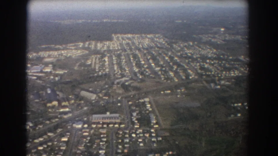 1967 Brisbane aircraft-window view showing suburban blocks, open ground, roadways, and scattered larger buildings.
